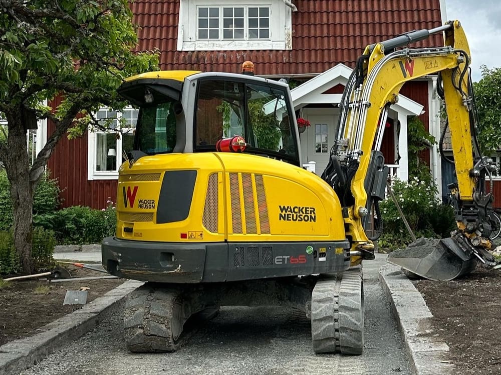 Grävmaskin förbereder en yta inför stenläggning vid ett rött hus