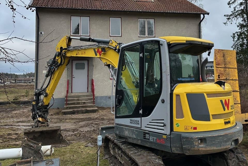 Grävmaskin från MM Gräv & Schakt framför ett bostadshus där husgrunden ska dräneras