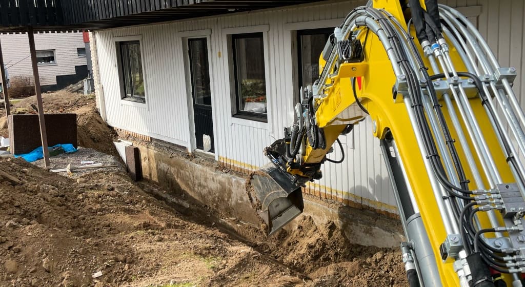 Grävmaskin schaktar längs grunden till ett bostadshus inför dränering